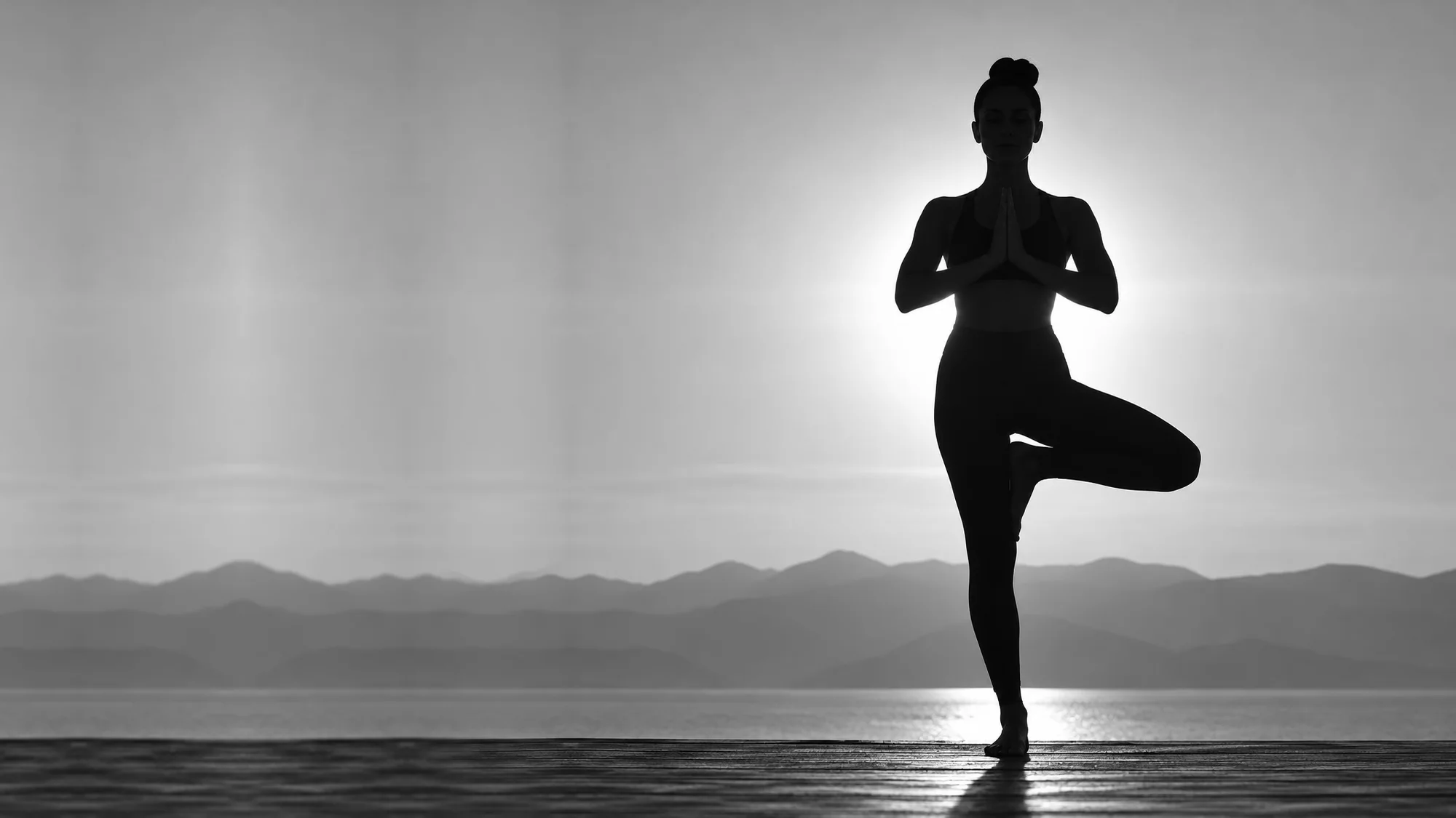 Silhouette of a woman in tree pose with hands at heart, sun and mountains behind her